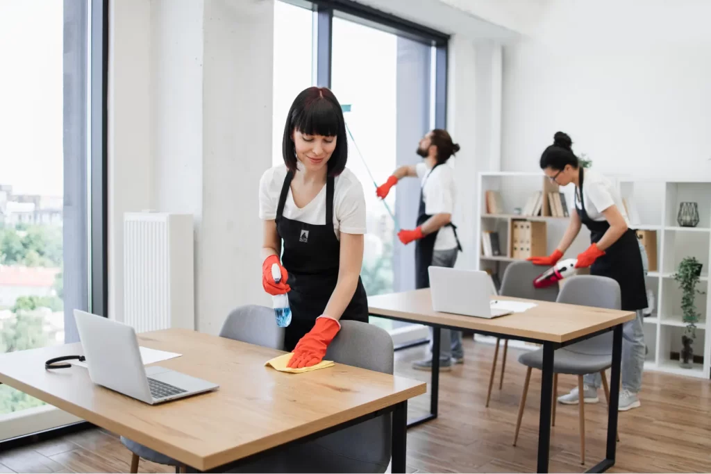 Female Elite Prime Clean cleaner wiping an office desk in a modern workspace with large windows, while a colleague cleans in the background