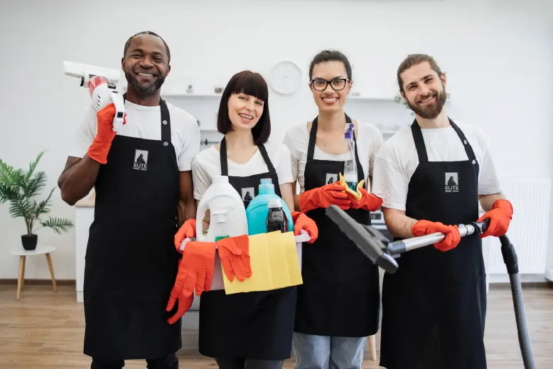 Four smiling Elite Prime Clean staff members wearing black aprons and holding cleaning supplies and a vacuum