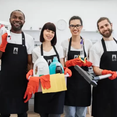 Four smiling Elite Prime Clean staff members wearing black aprons and holding cleaning supplies and a vacuum